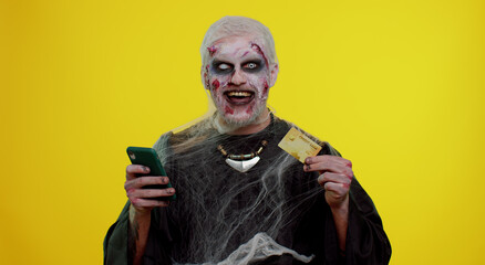 Sinister man with horrible scary Halloween zombie make-up using credit bank card and mobile phone, transferring money, purchases online shopping. Dead guy with wounded bloody scars face, yellow room