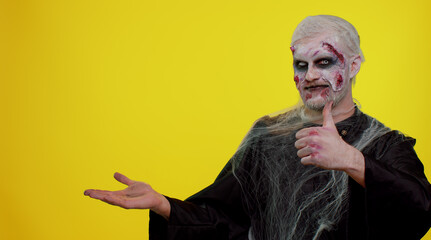 Zombie man with makeup with fake wounds scars showing thumbs up and pointing empty place,...