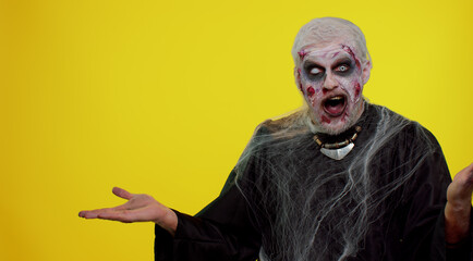 Creepy man with bloody scars face, Halloween stylish zombie make-up. Scary wounded undead guy showing thumbs up and pointing at left on blank space. Place for your advertisement logo. Sinister monster