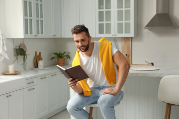Handsome man reading book on stool in kitchen