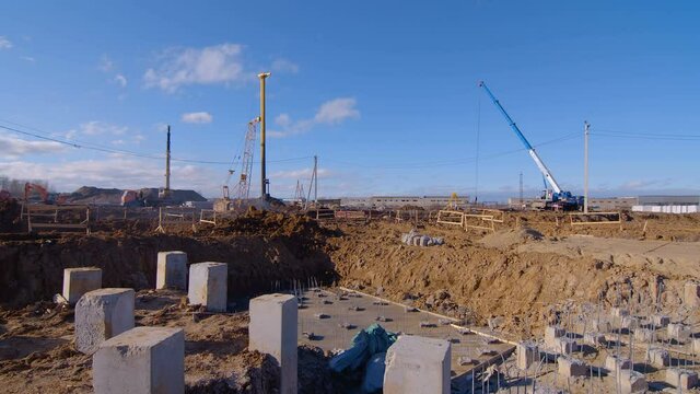 Сonstruction site at the stage of initial earthwork - an excavator, a pile driver, truck cranes are working. A foundation pit was dug, piles are now being driven into the ground 