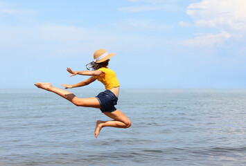 skinny girl with straw hat and yellow t-shirt performs high jump by the sea in summer