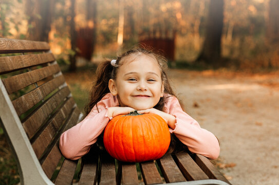 A little cheerful girl holds an orange pumpkin in the park in the autumn. Halloween props