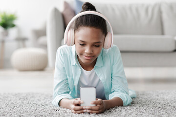 Concentrated smiling teen african american girl studying, watching online lesson on phone, lies on floor