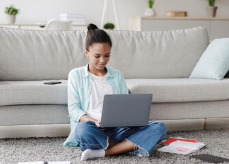 Busy concentrated teen afro american girl studying remotely, sitting on floor and look at laptop in living room