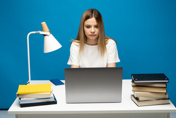 Portrait of a pretty young woman studying while sitting at the table with grey laptop computer, notebook. Smiling business woman working with a laptop isolated on a blue background.