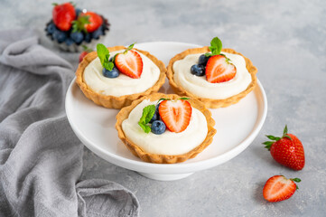 Delicious mini tarts with fresh berries and custard on a white plate on a gray concrete background. Copy space