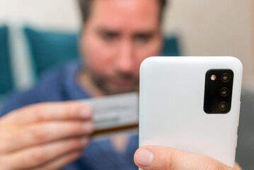 Young man buying fashion items through the Internet on cellphone by using credit card.Online Payment. Frontal portrait of man holding debit card and using smartphone for shopping at home