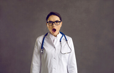 Studio shot of funny nurse or doctor surprised or scared by something. Young woman in glasses and white medical lab coat with stethoscope looking at camera with open mouth and shocked face expression