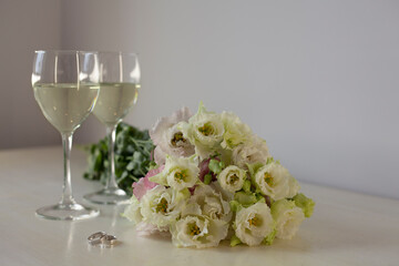 A bouquet of white flowers of eustom for wedding with two glasses of champagne and wedding rings. Classic wedding invitation with white copy space. Wedding ceremony accessories closeup.