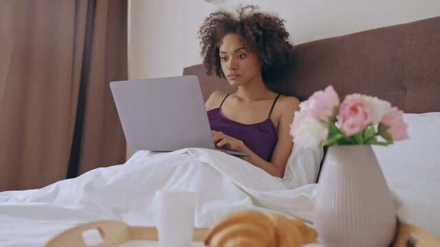 African-american woman working on laptop in bed, smiling, enjoying freelance job