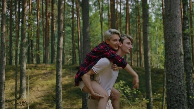 Young millennial couple spend time in nature, walk in green summer forest. Man carry woman in his arms. Piggy back ride of couple in love on outdoors date 