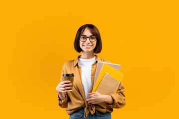 Happy female student holding notebooks and coffee to go, having break while studying, posing over yellow background