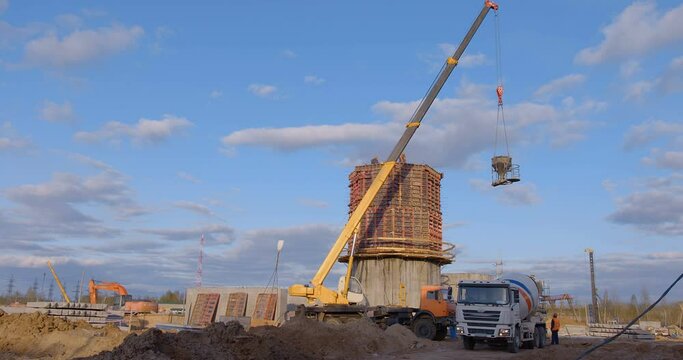 At construction site, a truck crane takes a bucket of cement from an autobenomixer and lifts it to the roof of a building under construction