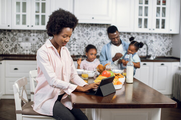 Focused woman working on digital tablet on bright kitchen while her husband feeding two daughters on background. African american family at home. Technology for lifestyles.