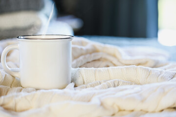 Fototapeta na wymiar Hot steaming cup of coffee sitting on top a soft white knit blanket on a bed with stack of covers in background. Selective focus with extreme blurred foreground and background. 