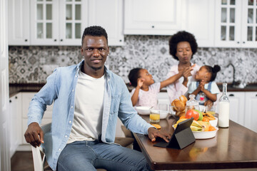 African american man sitting on bright kitchen with digital tablet and looking at camera. Blur background of charming wife playing with daughters during breakfast. Beautiful family at home.