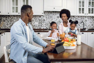 African american woman having breakfast with two cute daughters while husband working on digital tablet. Morning time of happy family on bright kitchen.