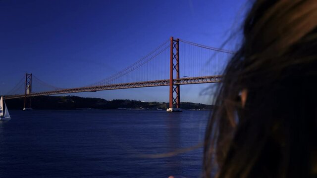 Woman taking photo on her smartpohne of the Ponte 25 de Abril bridge over the Tejo river in Lisbon at dusk - beautiful blue colors and calm waters