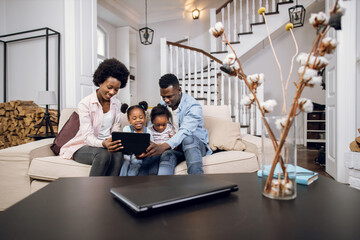African parents and their two cute daughters watching cartoons on digital tablet while resting together on comfy couch. Concept of family, technology and leisure time.