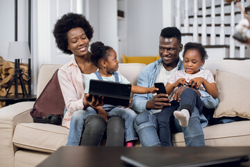 Cheerful african american family of four using joysticks and digital tablet for playing video games on comfy couch. Parents with two daughter spending time at home with fun.