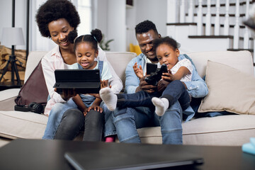 Happy african american family of four using joysticks and digital tablet for playing video games on comfy couch. Parents with two daughter spending time at home with fun.