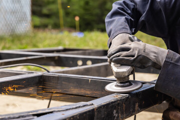 An image of a woman welder wearing protective gloves, running welding grinder on a metal surface, placed in a grassy field, in the daylight. 