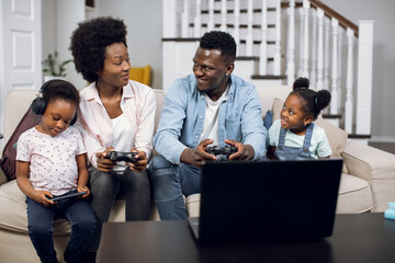 Smiling african man and woman sitting on sofa with wireless joysticks in hands and playing video games on laptop. Two cute daughters using smartphone and digital tablet.