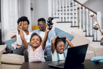 Cheerful african sisters playing video games on laptop with wireless joysticks while parents resting in hugs on couch. Concept of family, technology and lifestyles.