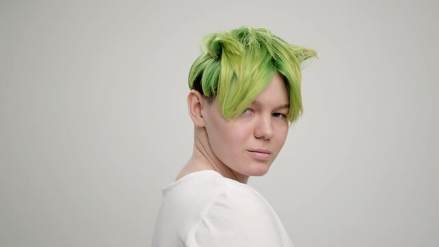 A young girl with a short pixie haircut and green hair in a white T-shirt on a light background. A woman poses in the studio, smiles and shows emotions. A breast portrait.