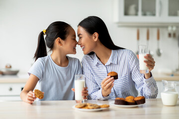Loving asian mother and little daughter having snacks and drinking milk in kitchen, enjoying...