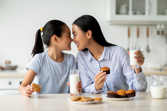 Loving asian mother and little daughter having snacks and drinking milk in kitchen, enjoying freshly baked cakes