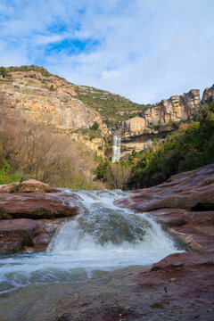 small waterfall between red rocks with cliff in the background with monastery, sant miquel del fai catalonia, spain