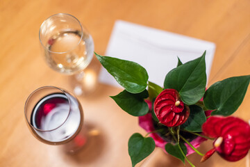 A beautiful view of a valentine set up on a wooden table, with red flowers and green leaves, beside a valentine note and two glasses of wine.