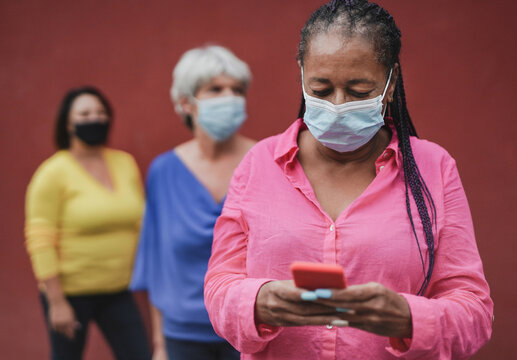 Multiracial women waiting in a line while wearing safety face maks for coronvirus - Elderly african woman using smartphone