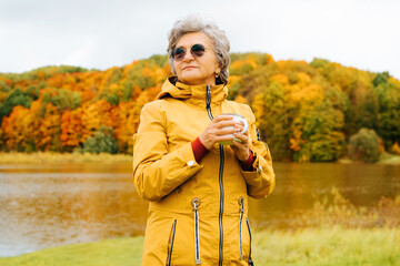 Portrait of attractive senior woman in glasses stands with mug hot drink, enjoys autumn against of lake and forest. Smiling retired woman in yellow jacket looking away outdoors. Retirement active