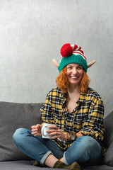 Portrait of a laughing red-haired girl in elf hat with mug in her hands. New Years mood. Vertical...