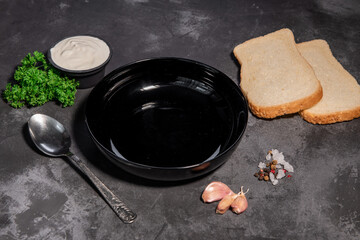 Table setting. Black soup plate on a black background. Spoon, white bread, garlic, spices, parsley, sour cream. Close-up