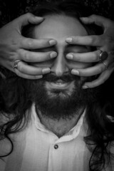 Black and white portrait of a young man with beard and long hair with eyes and face covered by a woman's hands