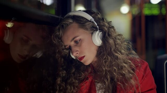 A depressed woman in headphones is riding in bus at night. Sad girl passenger is looking out window in modern tram. Depression or loss and problems or illness concept. City lights background.