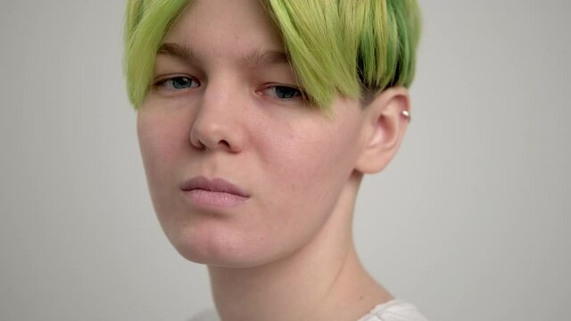 A young girl with a short pixie haircut and green hair in a white T-shirt on a light background. A woman poses in the studio, smiles and shows emotions. A breast portrait.