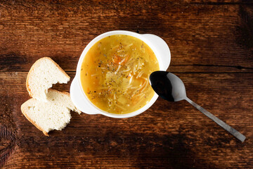 Chicken soup with noodles, potatoes, onions and carrots in a white deep plate with a black spoon and white bread on a wooden background.