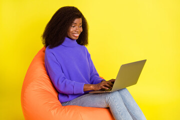 Profile portrait of cheerful dark skin lady sit soft chair use laptop beaming smile isolated on yellow color background