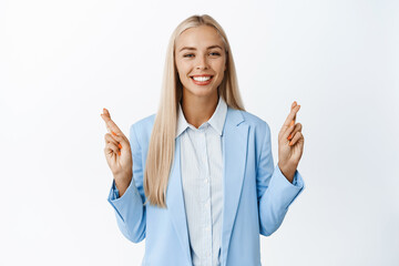 Hopeful smiling girl wishing, cross fingers for good luck, anticipating good news, standing in blue suit over white background