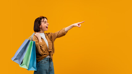 Wow, look at it. Excited lady pointing finger at free space and carrying colorful shopping bags, yellow background