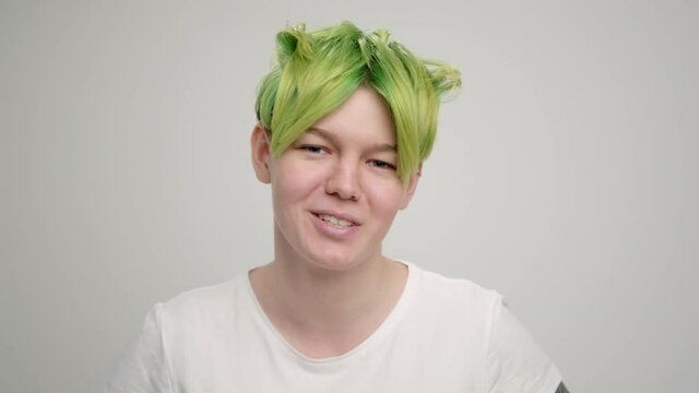 A young girl with a short pixie haircut and green hair in a white T-shirt on a light background. A woman poses in the studio, smiles and shows emotions. A breast portrait.