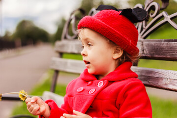 A cute little girl wearing red coat and a red hat sits on the wooden bench close up. Colorful city scene, copy space