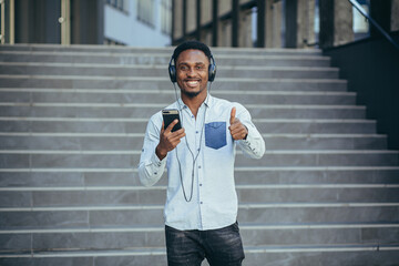 young african student listening to music from smartphone using big headphones, smiling from the...