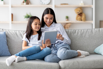 Happy young asian mom and daughter using digital tablet, watching videos or surfing internet,...