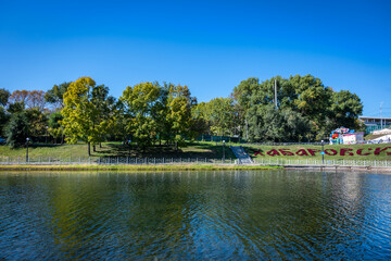 Fototapeta premium An autumn view of the public park in Khabarovsk (Russia) with blue clear sky, green and yellow trees, grass and a pond. On the grass there is an inscription 
